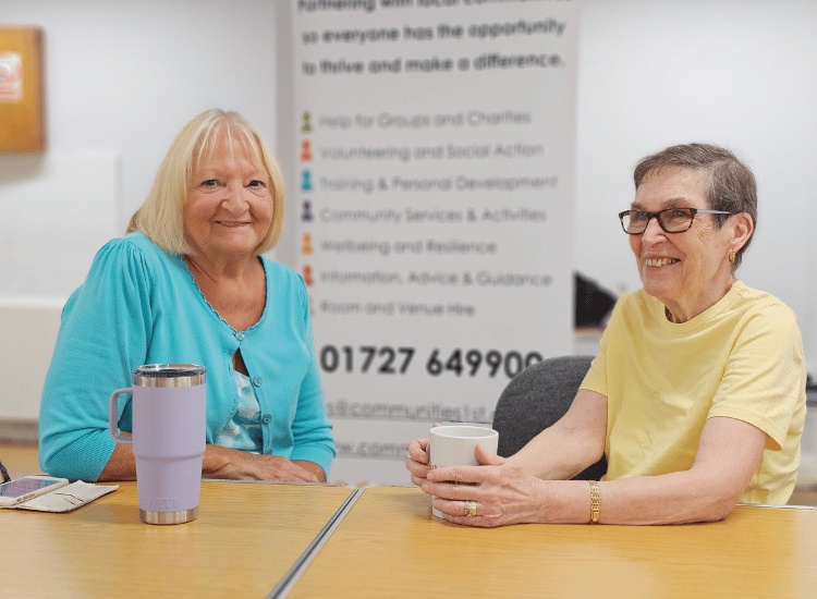 Two older women smiling and chatting over drinks, with a background banner listing Communities 1st services like 'Advice & Guidance' and 'Wellbeing and Resilience'.