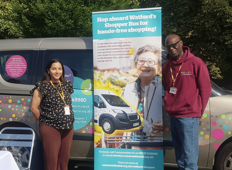 Two Communities 1st staff members standing next to a banner promoting Watford's Shopper Bus service, showing support for independent living.