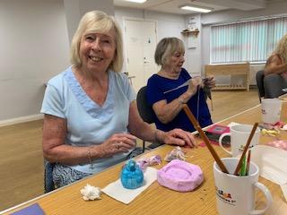 Smiling lady painting a small clay sculpture. The lady sitting next to her is knitting