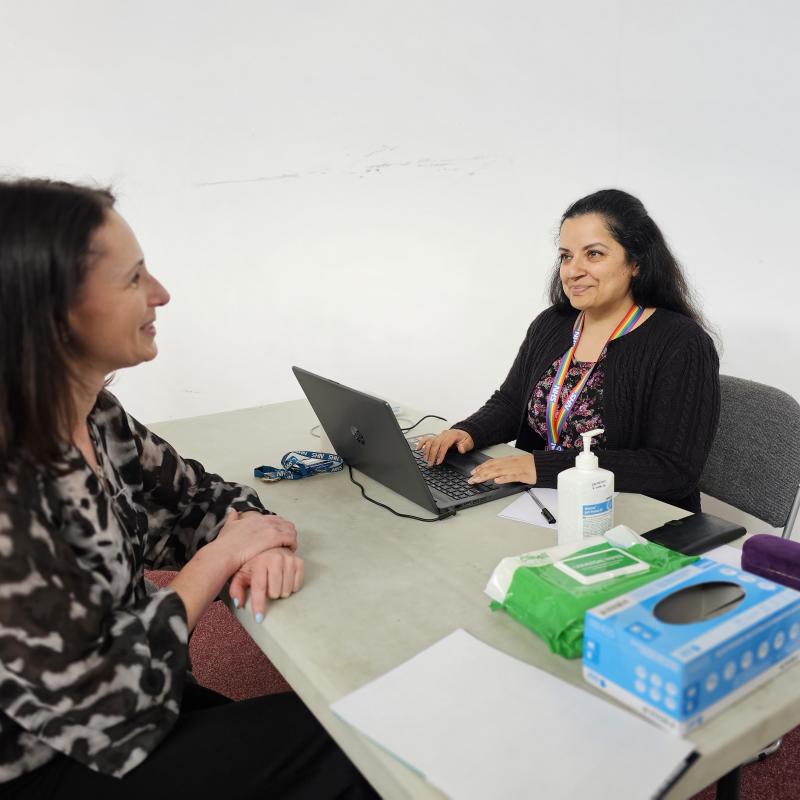 A woman giving support to another using a laptop, facing across the table from each other.